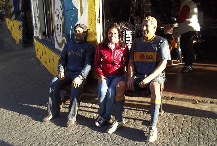 Girl wearing red coat sitting between two soccer players' statues.