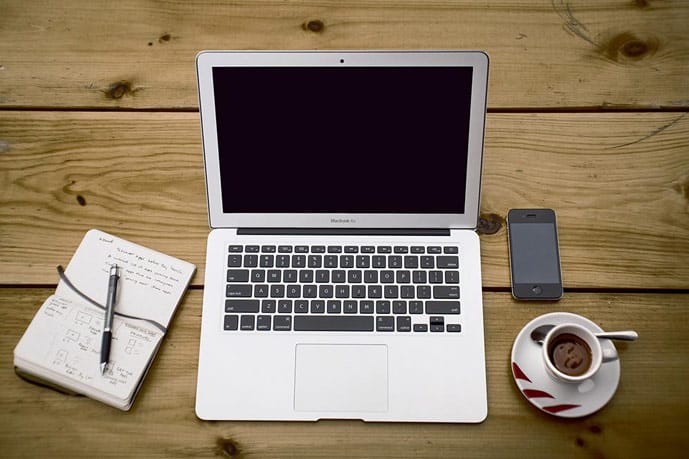 Laptop, notebook, cell phone, and a cup of coffee on a wooden table.