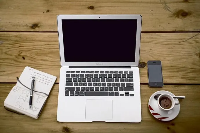 Laptop, notebook, cell phone, and a cup of coffee on a wooden table.