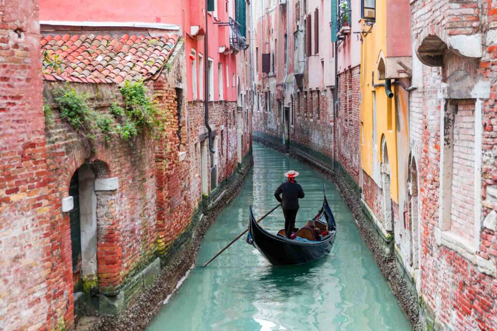 Venetian gondolier punting gondola through green canal waters of Venice, Italy